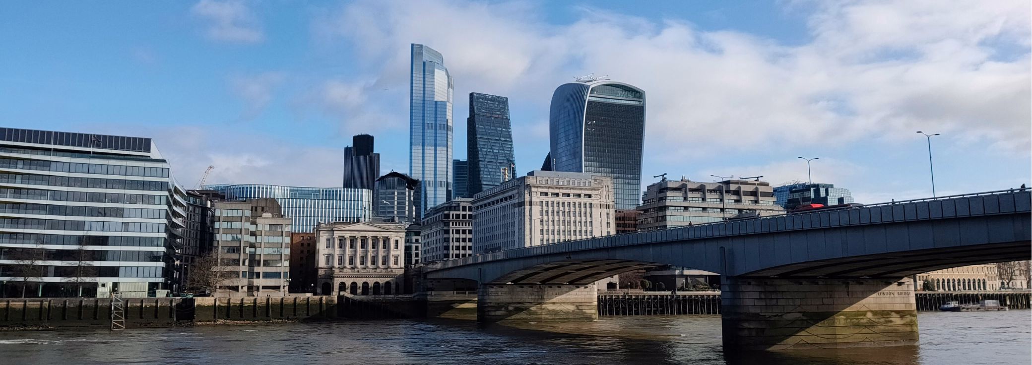 A view from the River Thames highlights London Bridge stretching toward a city skyline featuring the Walkie Talkie building, set under a blue sky.