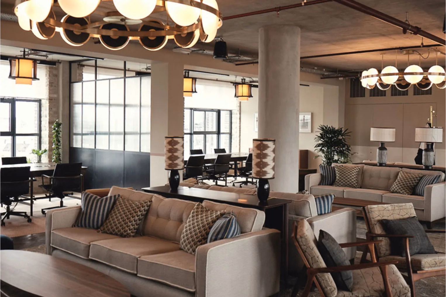 Beige sofas and wooden armchairs in a lounge area featuring circular ring chandeliers, concrete structural pillars, and a background office workspace.