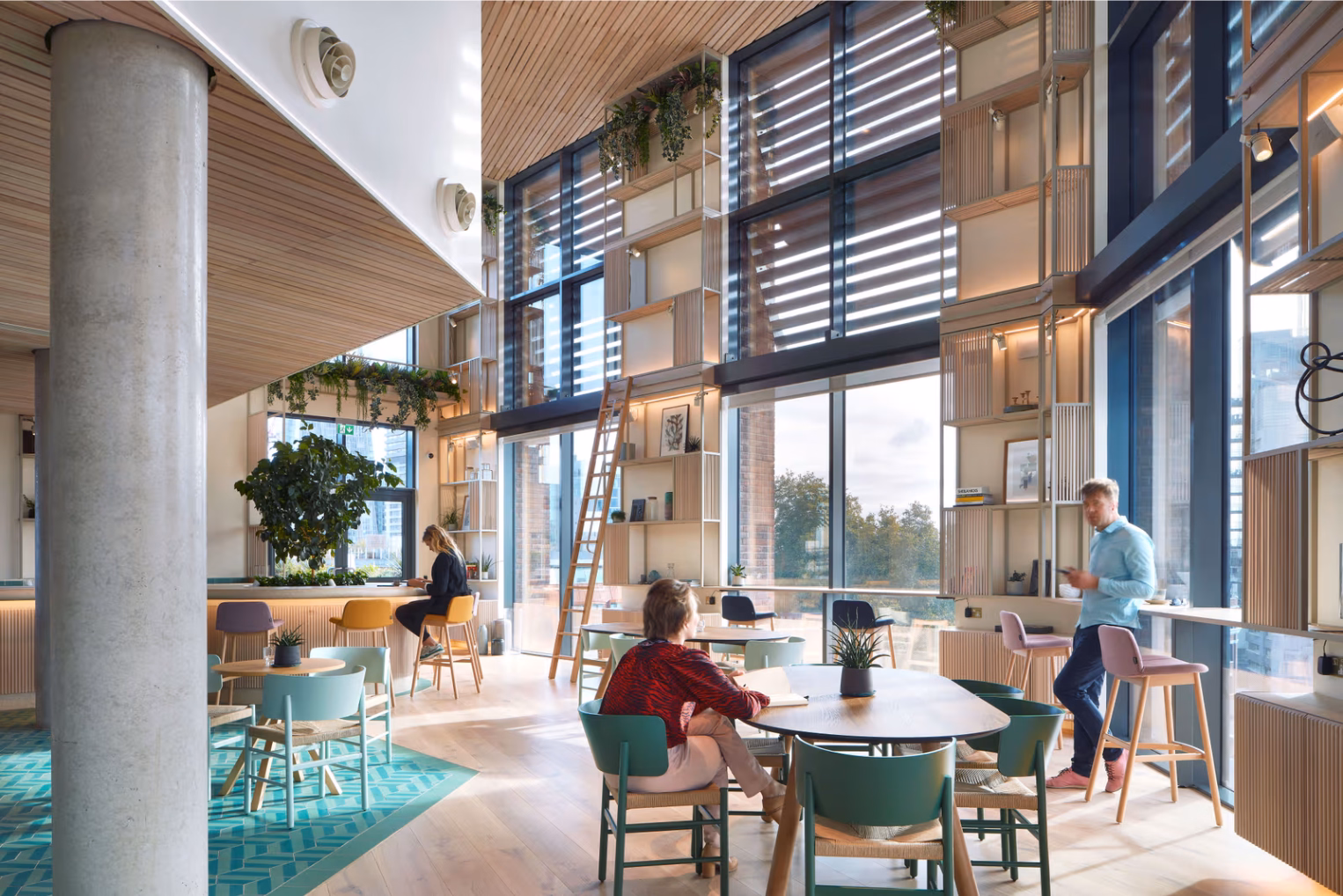 People sit and stand at wooden tables and counters with colourful chairs and teal patterned flooring, surrounded by wooden shelving units and floor-to-ceiling windows.