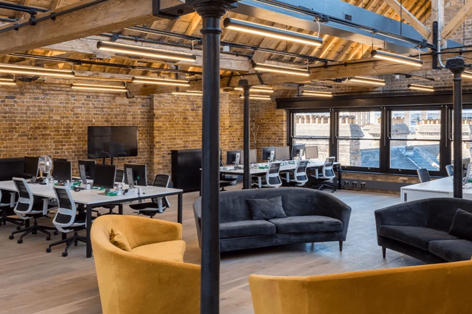 Yellow and grey sofas in a central lounge area surrounded by white office desks with white-and-black mesh chairs under an exposed brick wall.