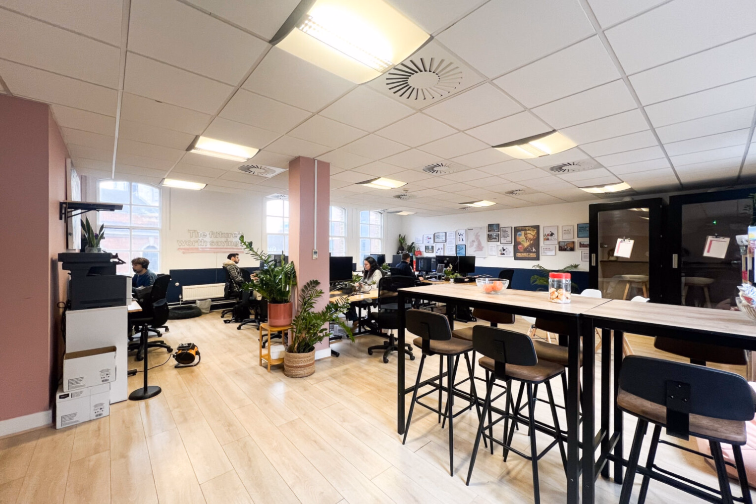 Barstools are under a high-top table, while several employees work at desks with black chairs in an open office space with a pink support column and a white grid ceiling.
