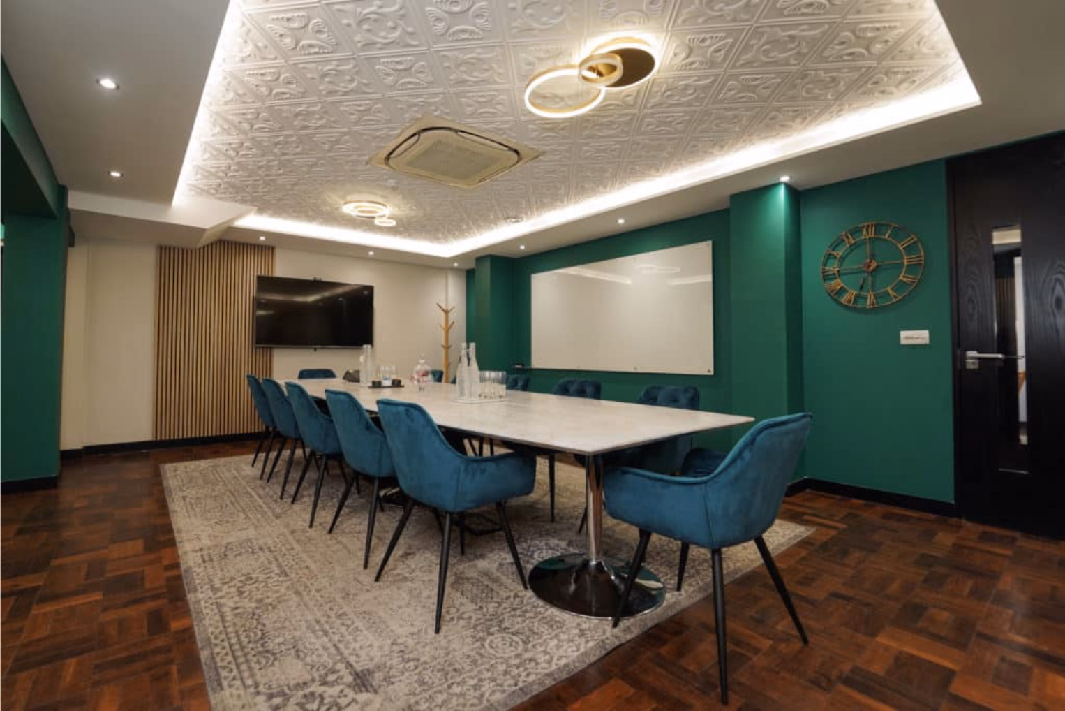 Blue velvet armchairs surround a long table on a patterned grey rug in a green room.