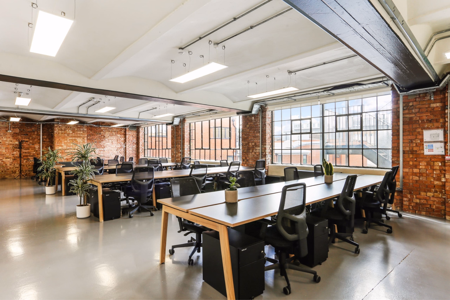 Black mesh chairs at wooden desks on a smooth grey floor featuring large multi-pane windows and exposed red brick walls.