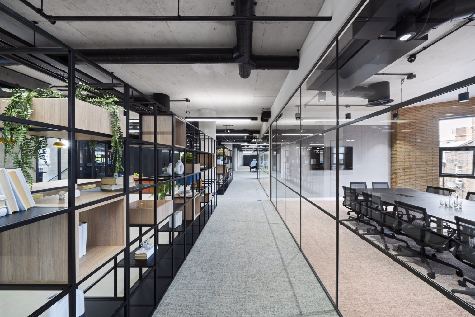 An office hallway with light grey carpeting, an open black metal shelving unit with wooden box inserts, and a glass-walled room with a dark table and mesh chairs.