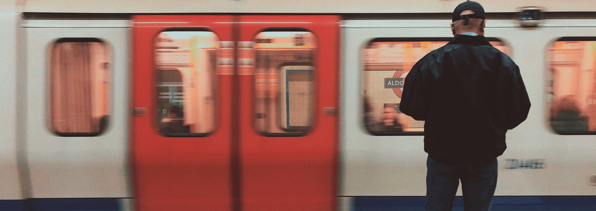 A man wearing a black jacket and a baseball cap on a platform from behind as a London Underground train drives past.