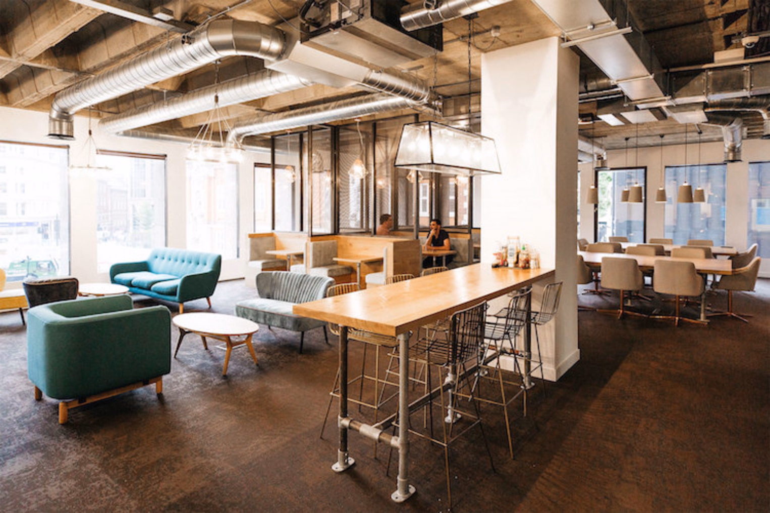 A wooden high-top table with metal wire barstools featuring a mix of green and grey sofas, a dining table, and dining booths under exposed industrial ductwork.
