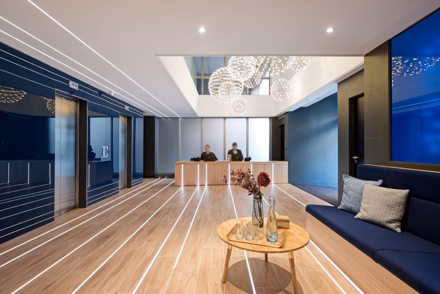 Glowing linear LED strip lights embedded in a hardwood floor lead toward a light wood reception desk, flanked by a blue bench, under spherical chandeliers.