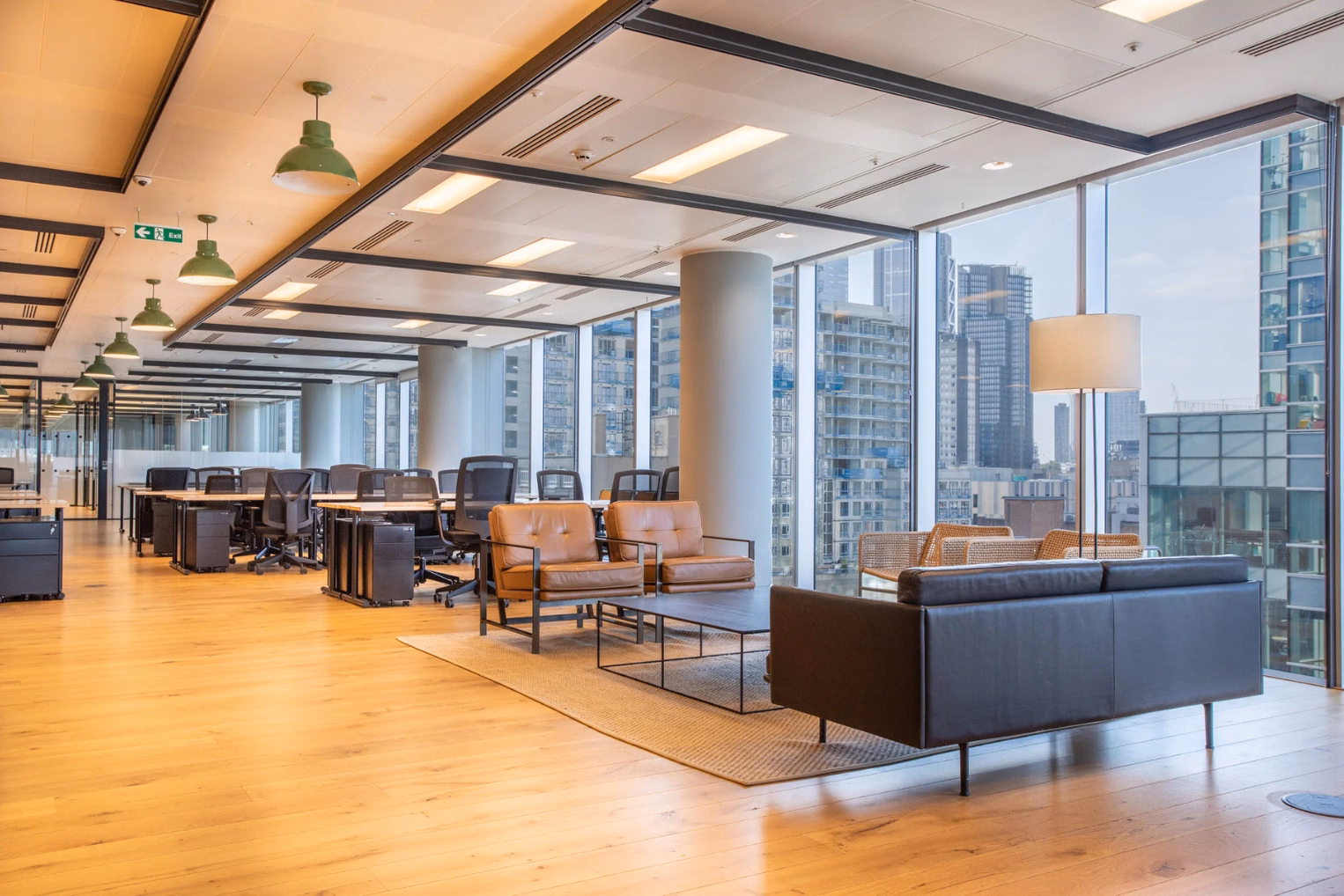 A black leather sofa and tan leather armchairs around a low coffee table on a neutral rug, next to floor-to-ceiling windows with a city skyline view and rows of office desks.