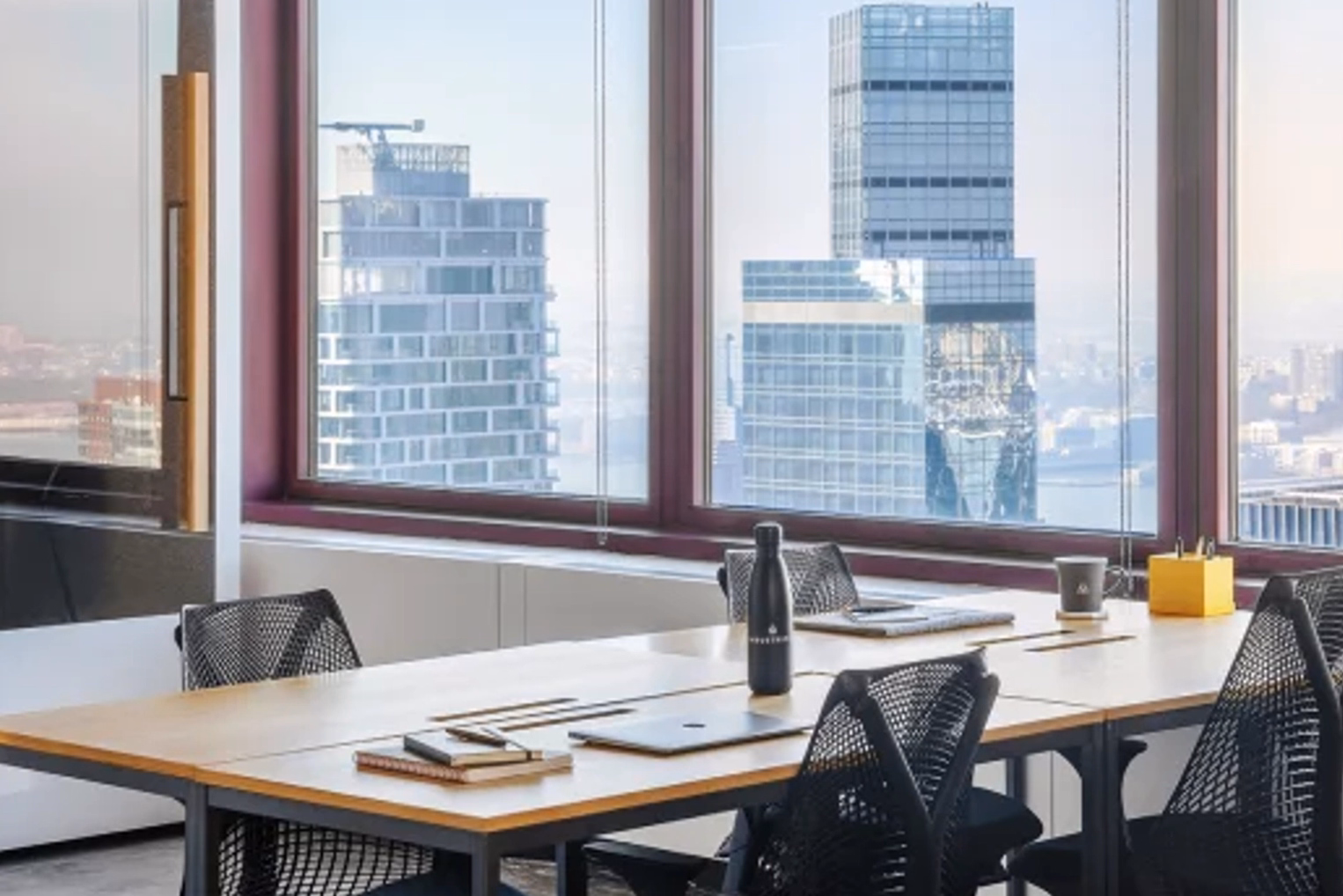 Black mesh office chairs surround a long wooden desk with notebooks and laptops, positioned next to large windows that look out on a towering city skyline.