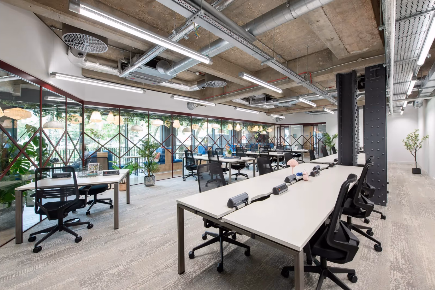 Black office chairs at white desks in a bright workspace featuring industrial piping on the ceiling and a glass wall partitioning.