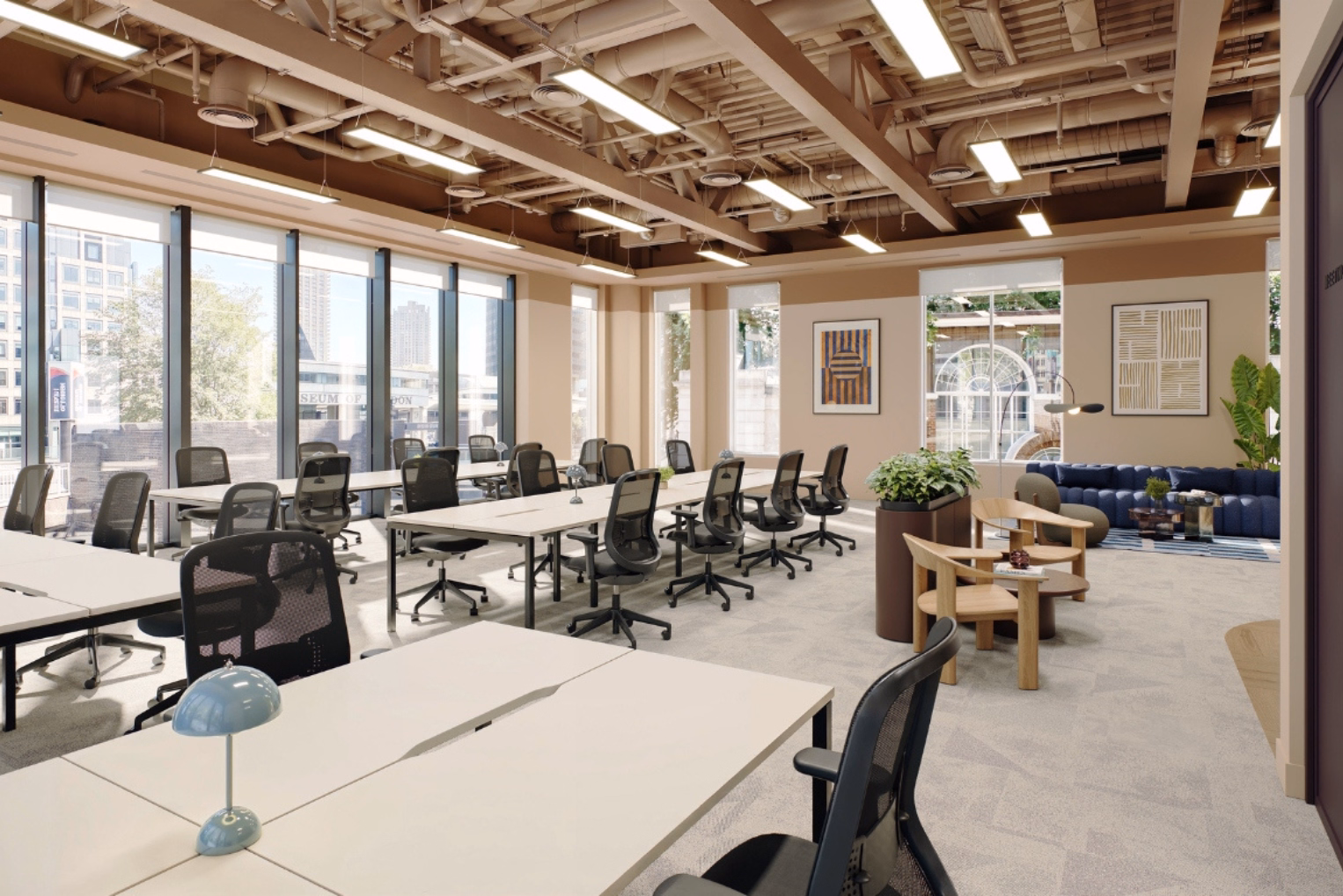 Black mesh chairs with white desks in an open-plan workspace, which includes wooden armchairs and a blue sofa under an exposed industrial ceiling with wooden beams.