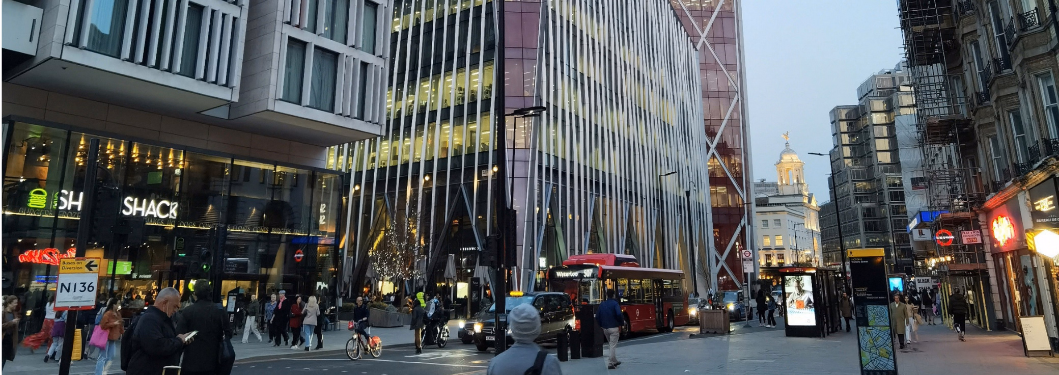 A busy city street features glass skyscrapers with pedestrians, a red bus, a black cab, and a Shake Shack restaurant on the corner.