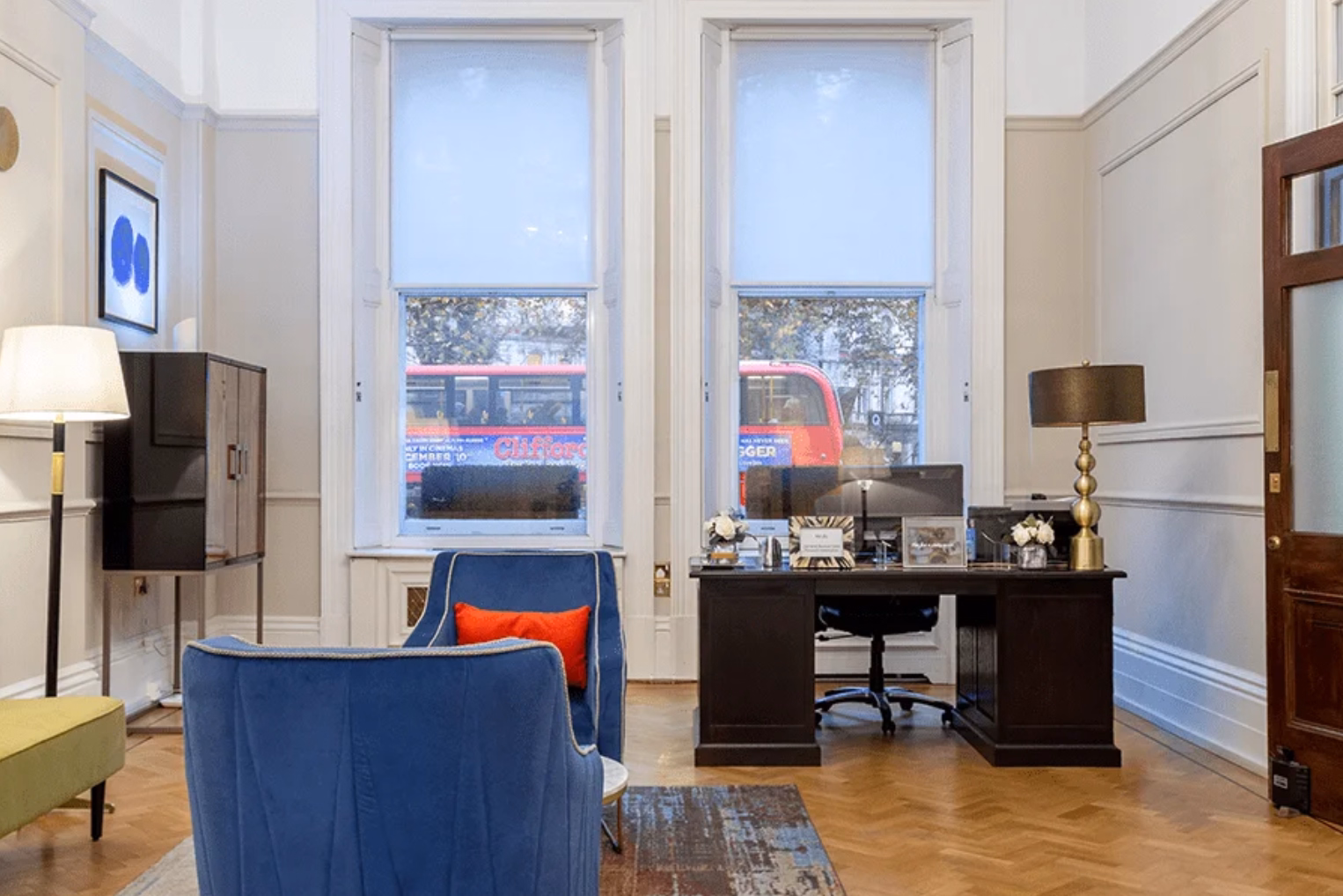 Blue velvet armchairs on a patterned rug in front of a wooden desk near large windows framing a red double-decker bus outside.