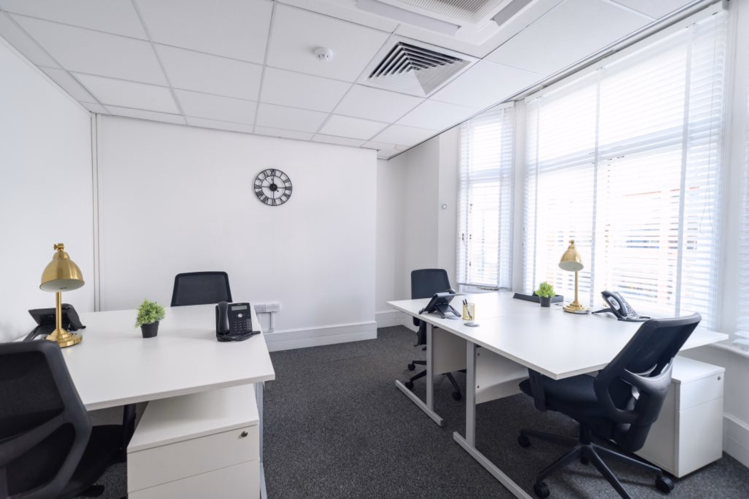Black mesh chairs face white desks with gold lamps and desktop phones on grey carpeted floor near large windows covered by white blinds.