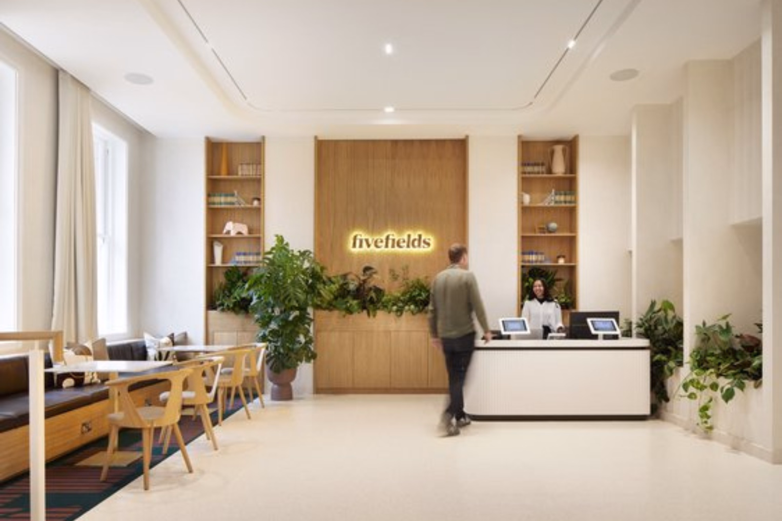 A white reception desk across from a seating area with wooden chairs and a bench, a wood-paneled wall with a "fivefields" sign and built-in bookshelves.