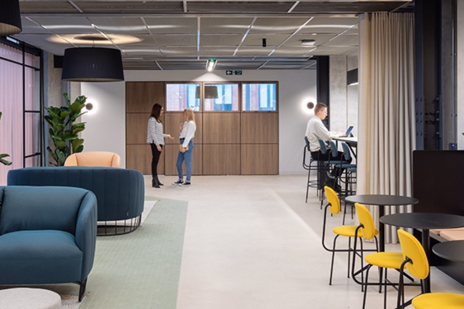 Yellow chairs around black café tables, plus teal armchairs on a green rug, a high-top counter workspace with barstools, and a wood-panelled wall in the background.