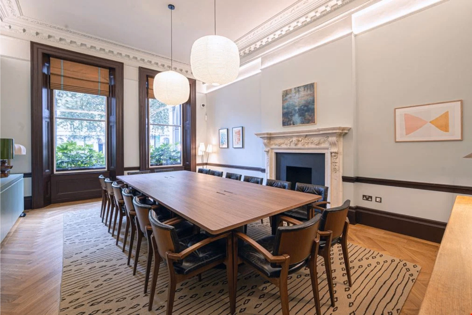 A wooden table surrounded by black leather and wood armchairs on a patterned cream rug in a meeting room featuring a white fireplace with two large windows.