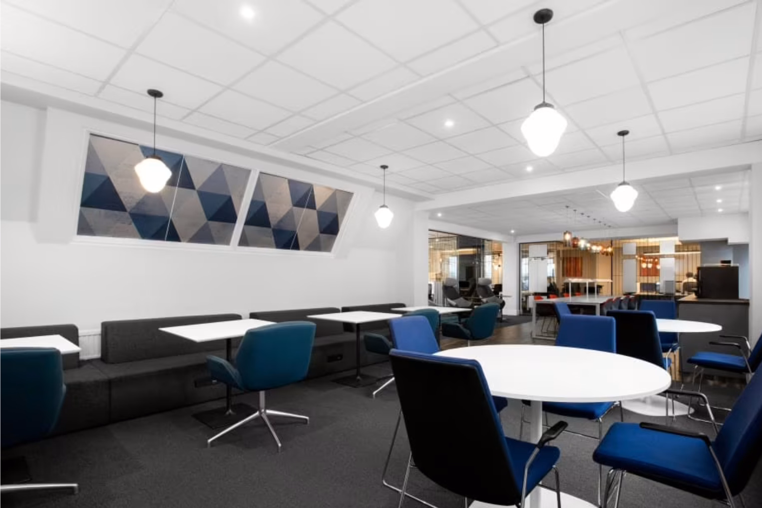 A communal area featuring white round and square tables paired with blue chairs, and a grey bench sits below a geometric panelled window.