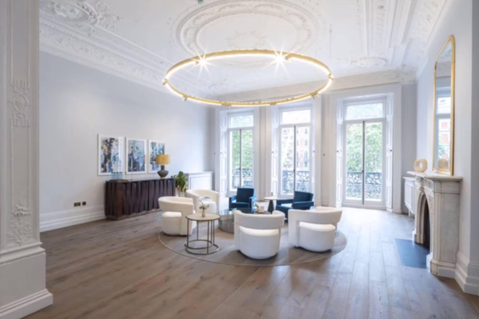 White bucket chairs and blue armchairs on a round rug, illuminated by a large, glowing circular halo chandelier.