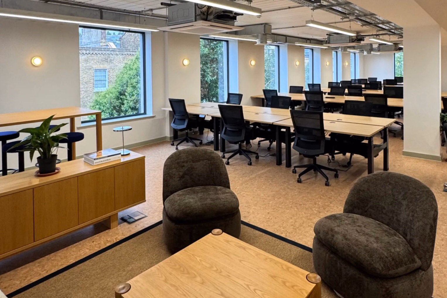 Two charcoal armchairs and a wooden table in an open-plan office, which features multiple rows of wooden desks with black mesh chairs.