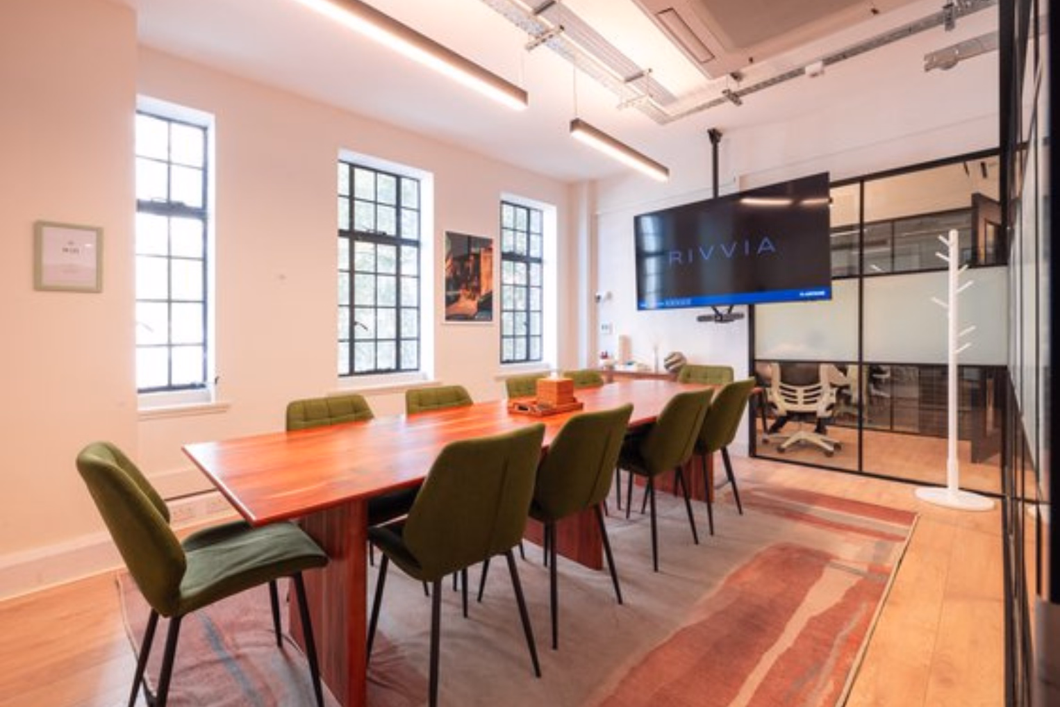 Green chairs surround a long wooden table on an abstract patterned rug, under tube pendant lighting in a meeting room.