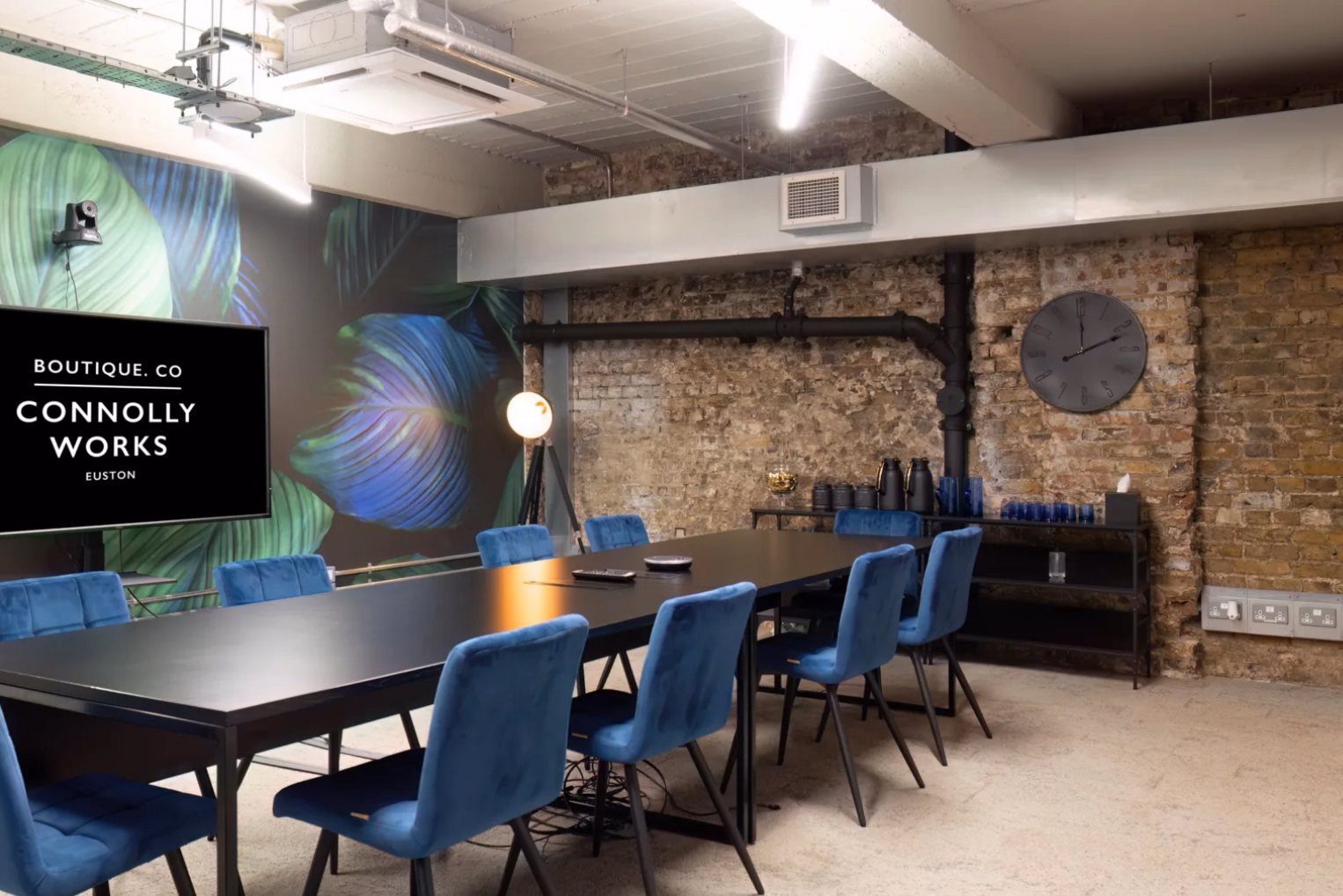 Blue velvet chairs surround a black conference table in an industrial-style meeting room featuring an exposed brick wall and a large display screen.