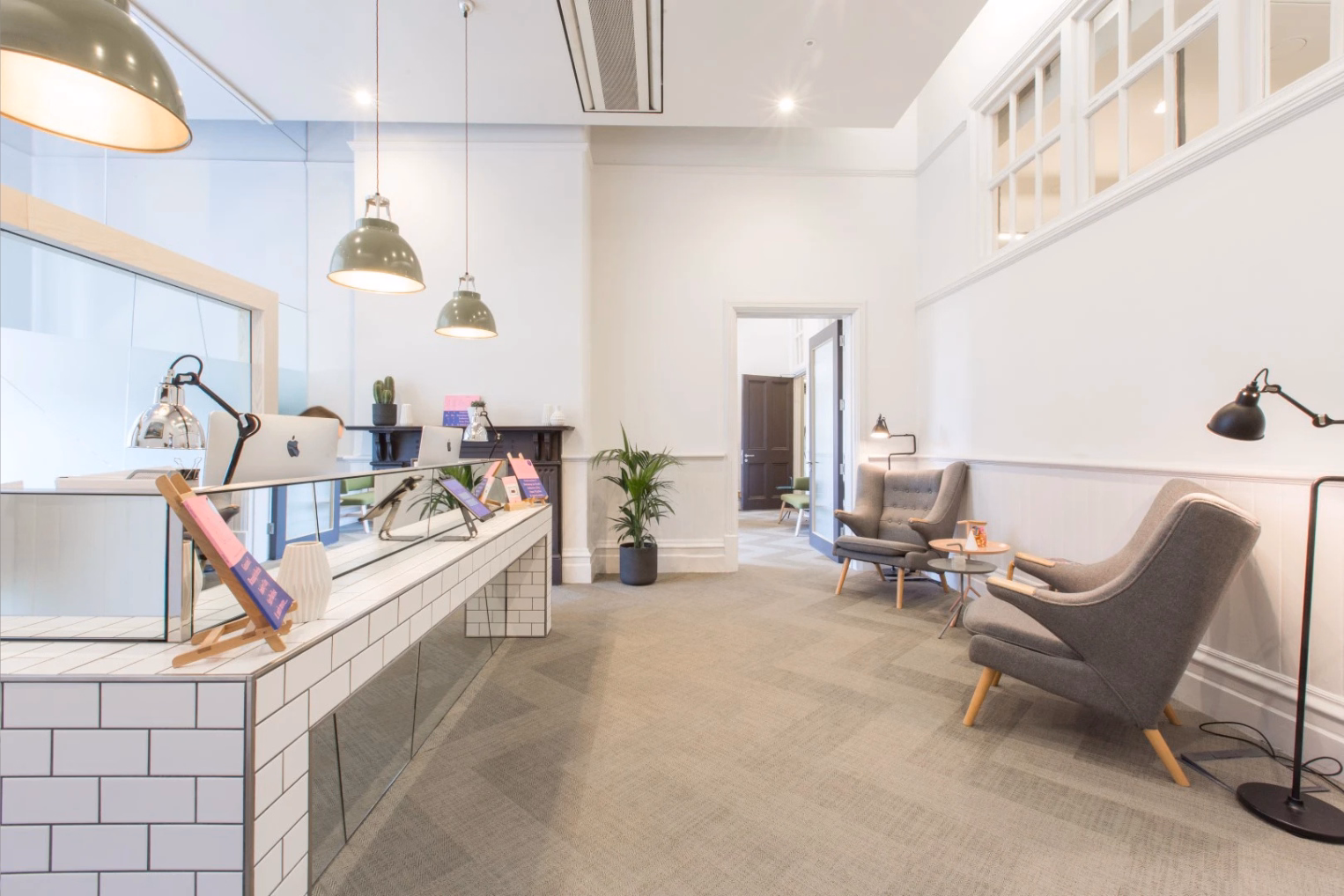 Two grey armchairs sit in a waiting area across from a white-tiled reception desk, with high ceilings and green pendant lights.