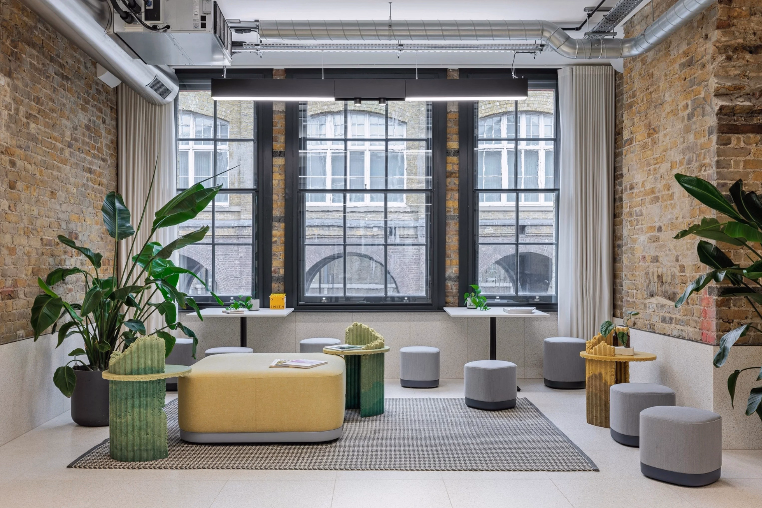  A yellow ottoman on a patterned grey rug, green accent tables, green potted plants, small grey stools, and three large black-framed windows.