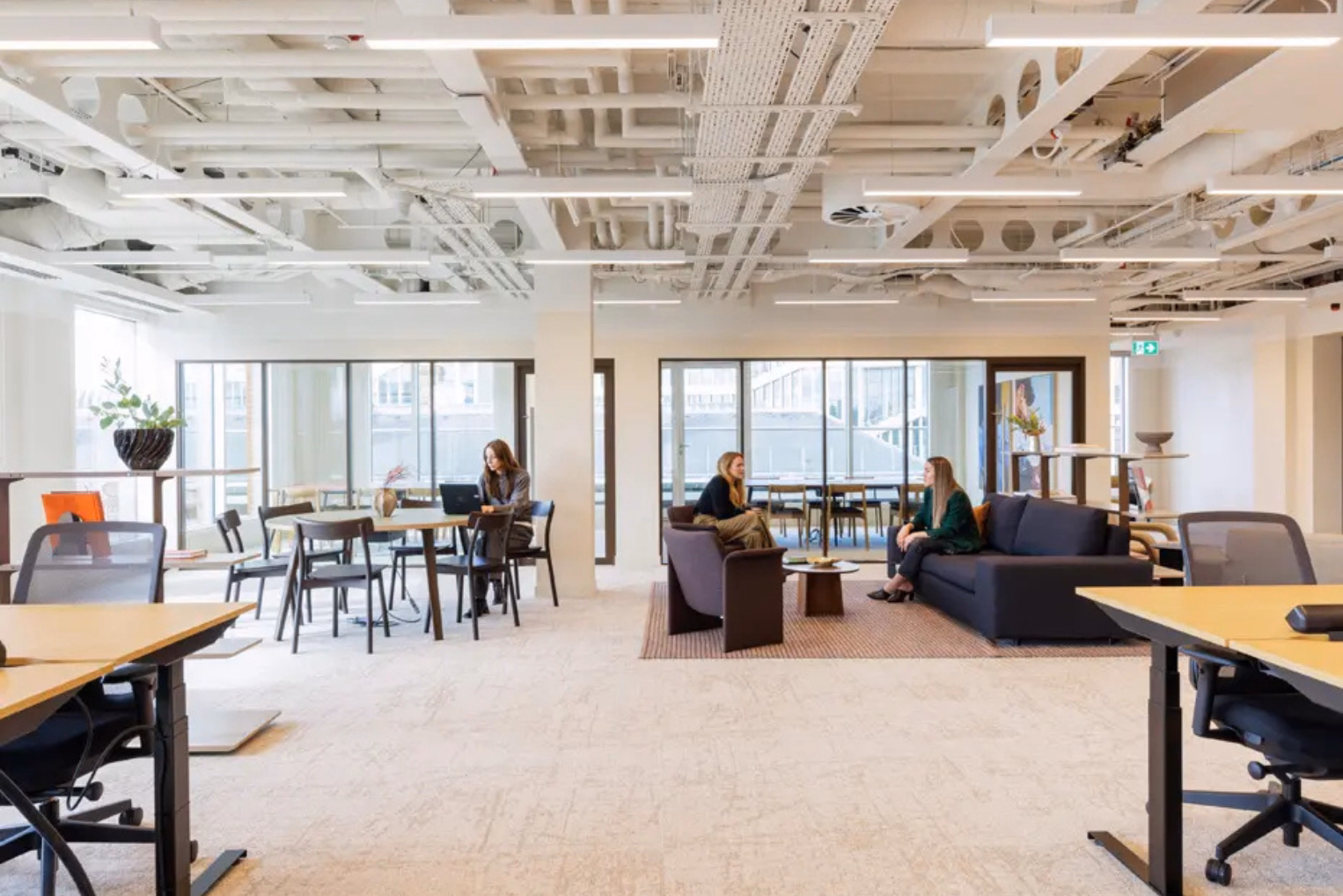 People sit in a lounge between rows of wooden desks with mesh office chairs.