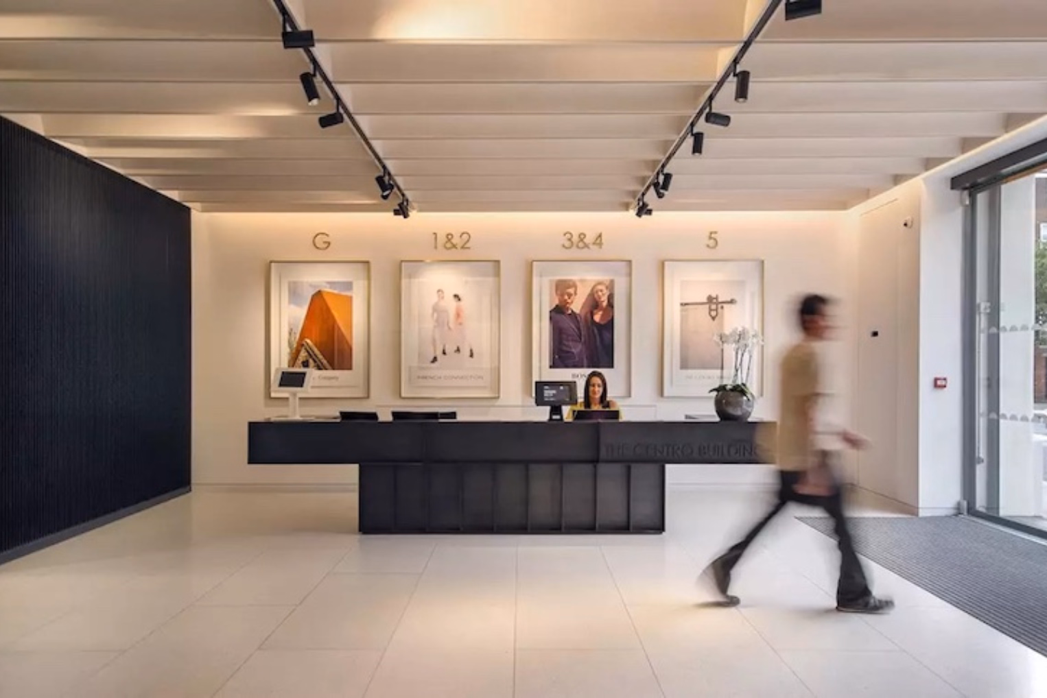  A dark reception desk sits on a white tiled floor, with four large framed prints with gold floor labels under a slatted ceiling with black track lighting.