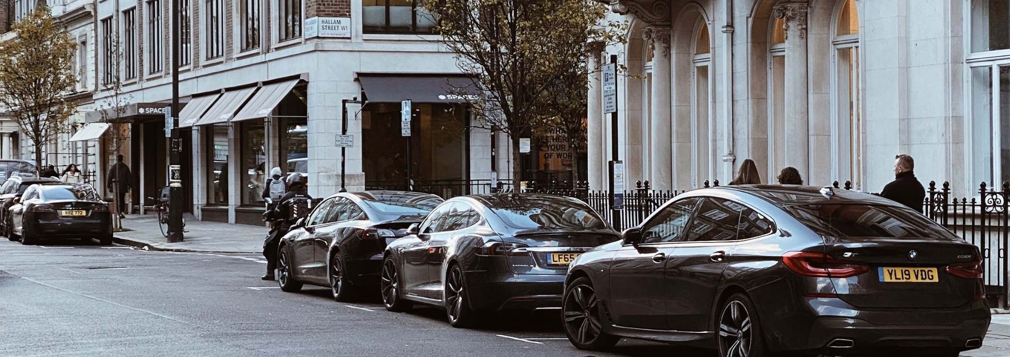 Several cars parked along the side of a city street lined with classic stone buildings, storefronts with dark awnings, and thin trees.