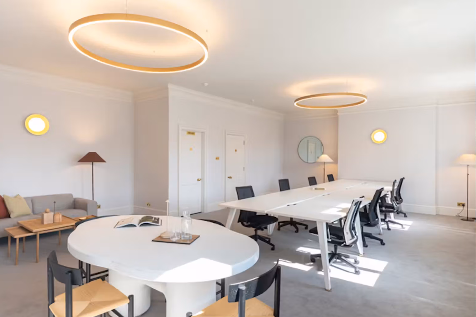 An open-plan office features a long white table with black rolling chairs alongside a round white meeting table with wooden chairs, illuminated by two large halo chandeliers.