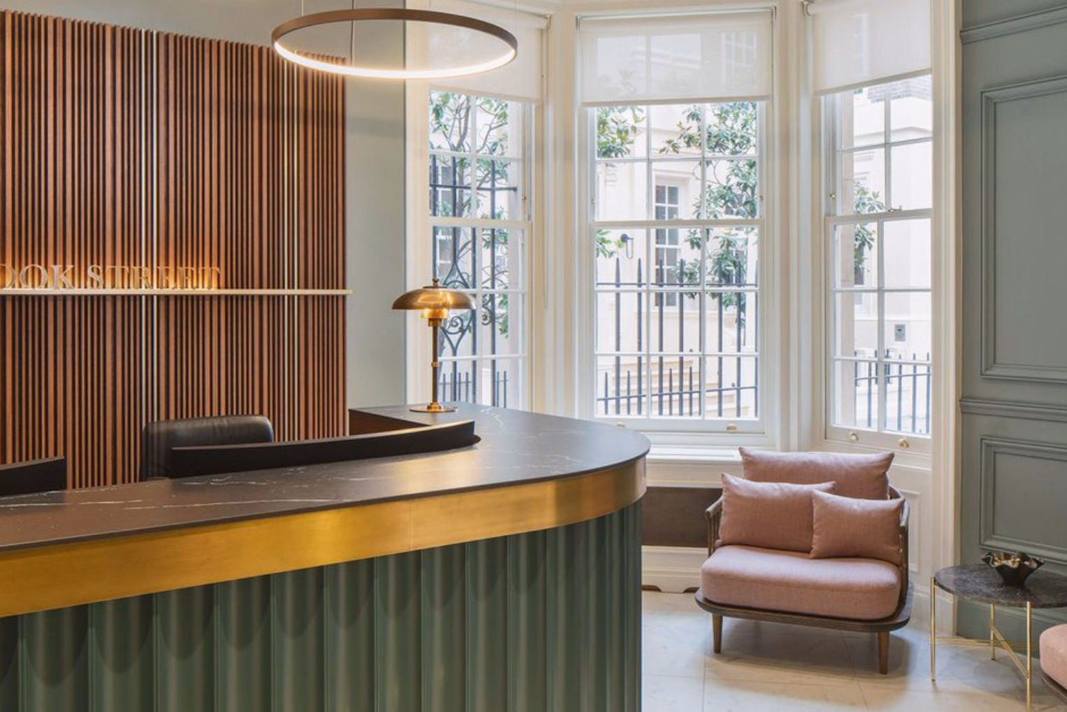 A curved, green fluted reception desk with a gold trim and dark countertop stands in a waiting area, complemented by a dusty pink armchair nestled in a large bay window.