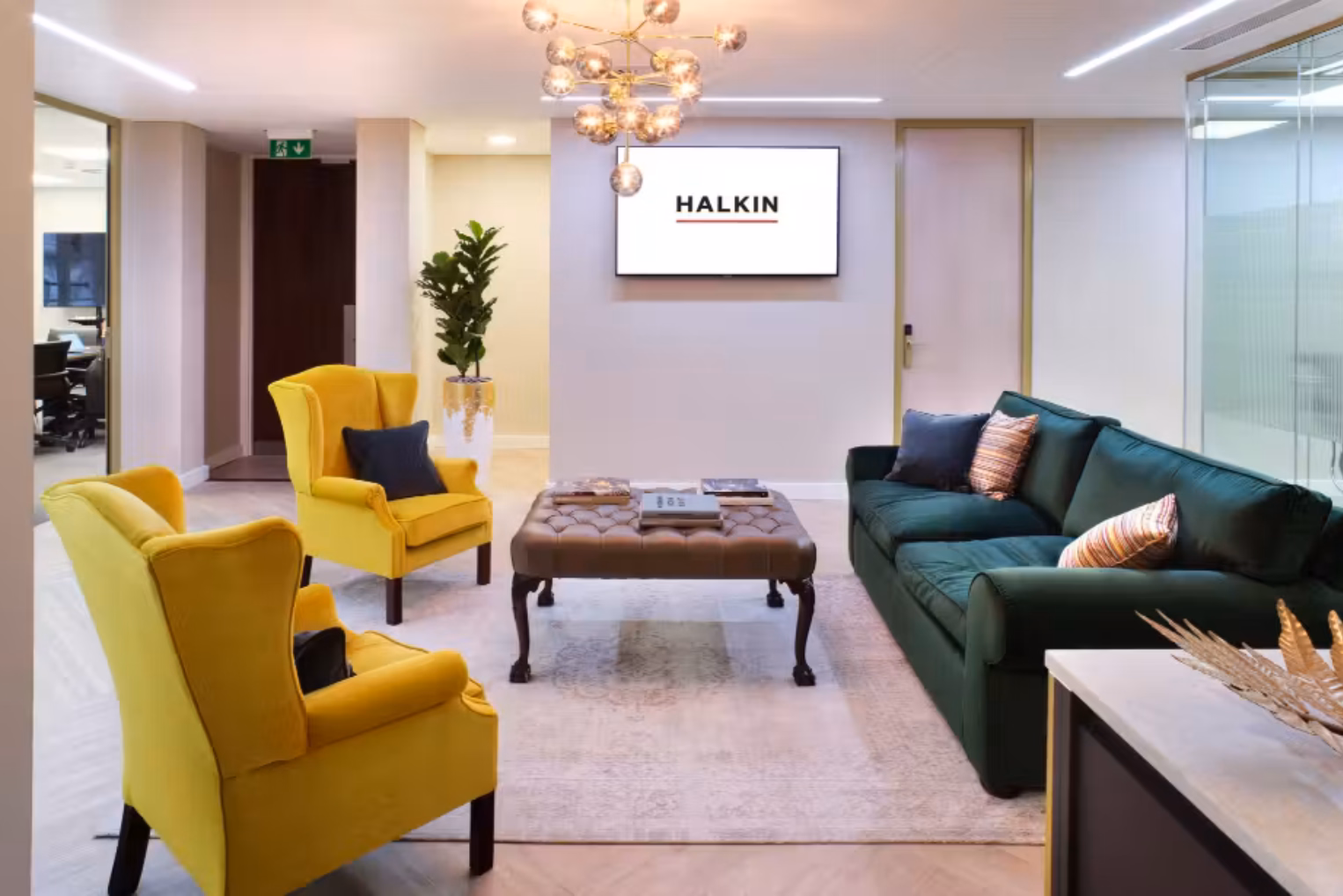 A waiting area features two yellow armchairs facing a dark green velvet sofa, around a brown leather table under a bubble chandelier and a wall-mounted screen.