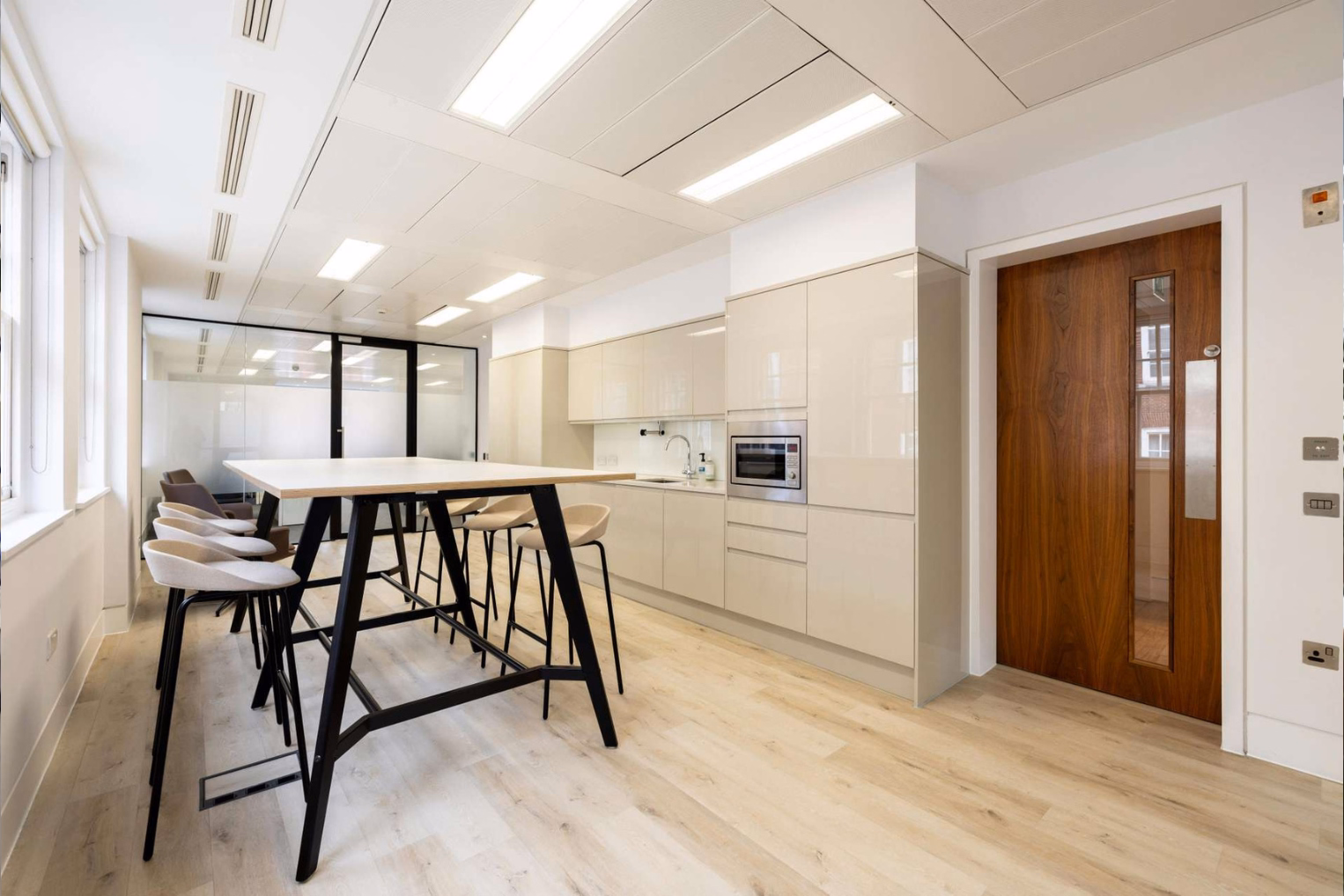 A kitchen and break area features cream-coloured cabinets with a built-in microwave alongside a high communal table with light grey barstools.