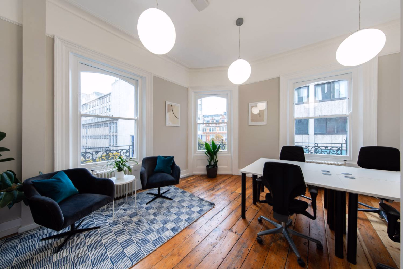 An office space features a white desk with black rolling chairs opposite a small seating area with two armchairs and teal cushions on a blue checkered rug.