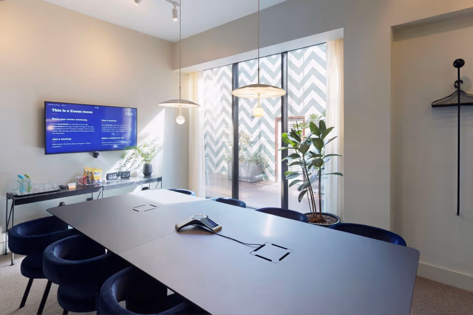 Blue velvet chairs surround a grey table in a meeting room, featuring low-hanging lights and a wall-mounted screen next to glass doors that open to an outdoor patio.