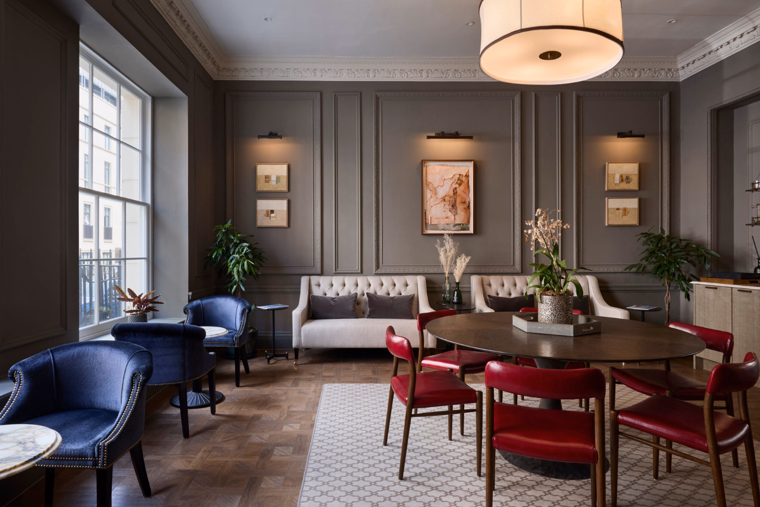 Red leather chairs surround a wooden dining table on a patterned cream rug opposite blue velvet club chairs and two beige sofas.