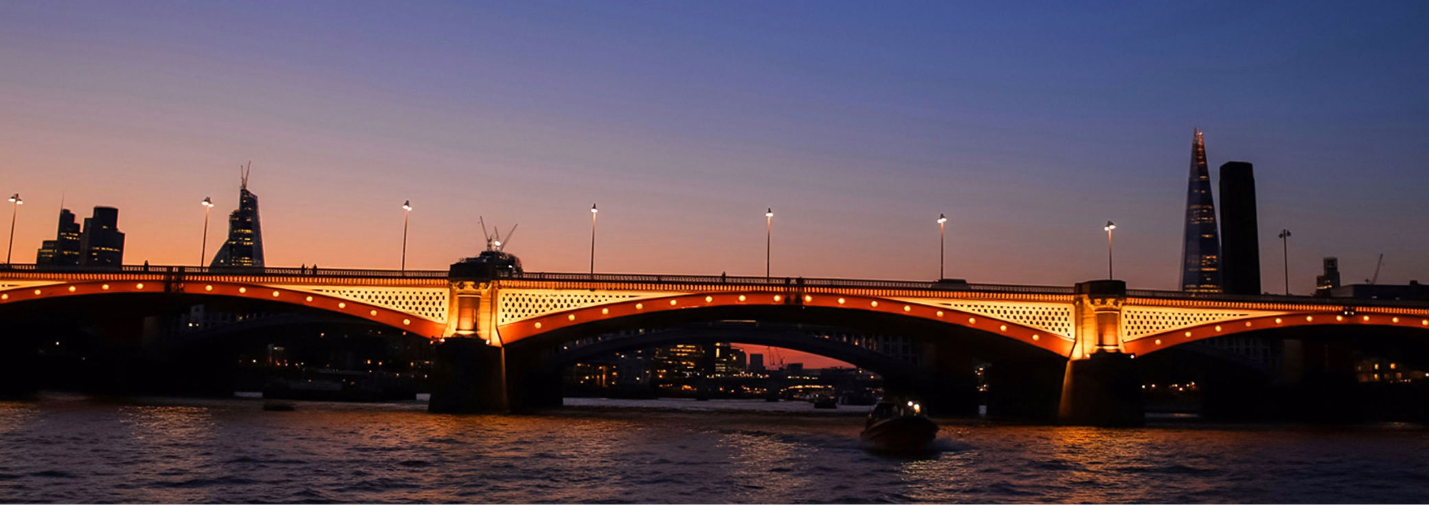An illuminated arched bridge spans a wide river, casting an orange glow onto the water as a city skyline, including The Shard, silhouettes against a sunset sky.
