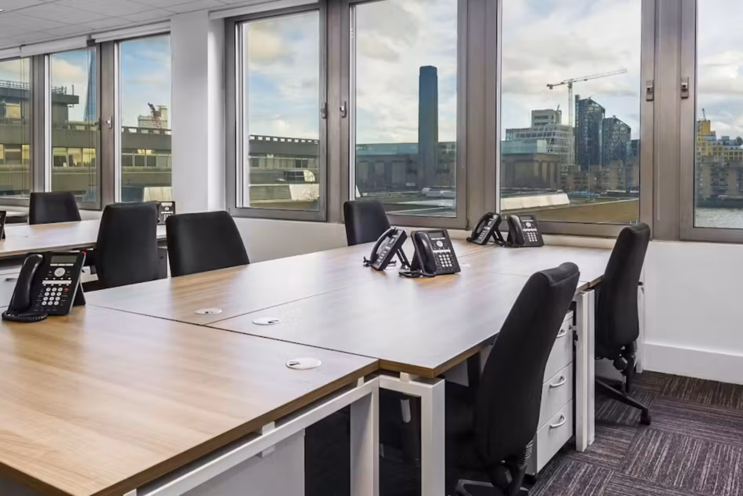 Black chairs and desk phones on light wooden desks in a bright workspace, featuring large windows overlooking a city skyline with the Tate Modern.