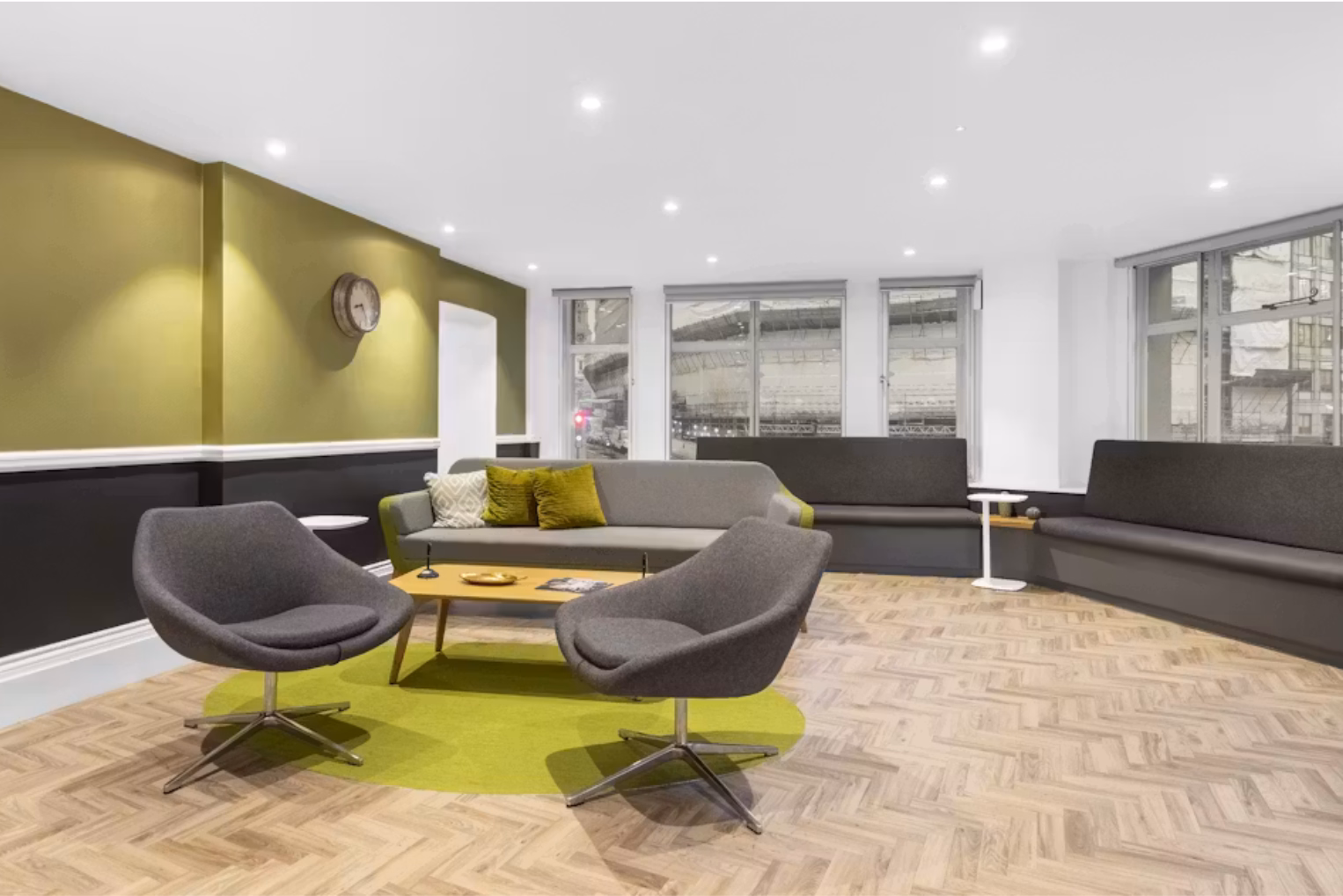 Two dark grey armchairs sit on a lime-green rug in an office lounge, facing a grey sofa with green pillows, with built-in dark grey banquette seating.