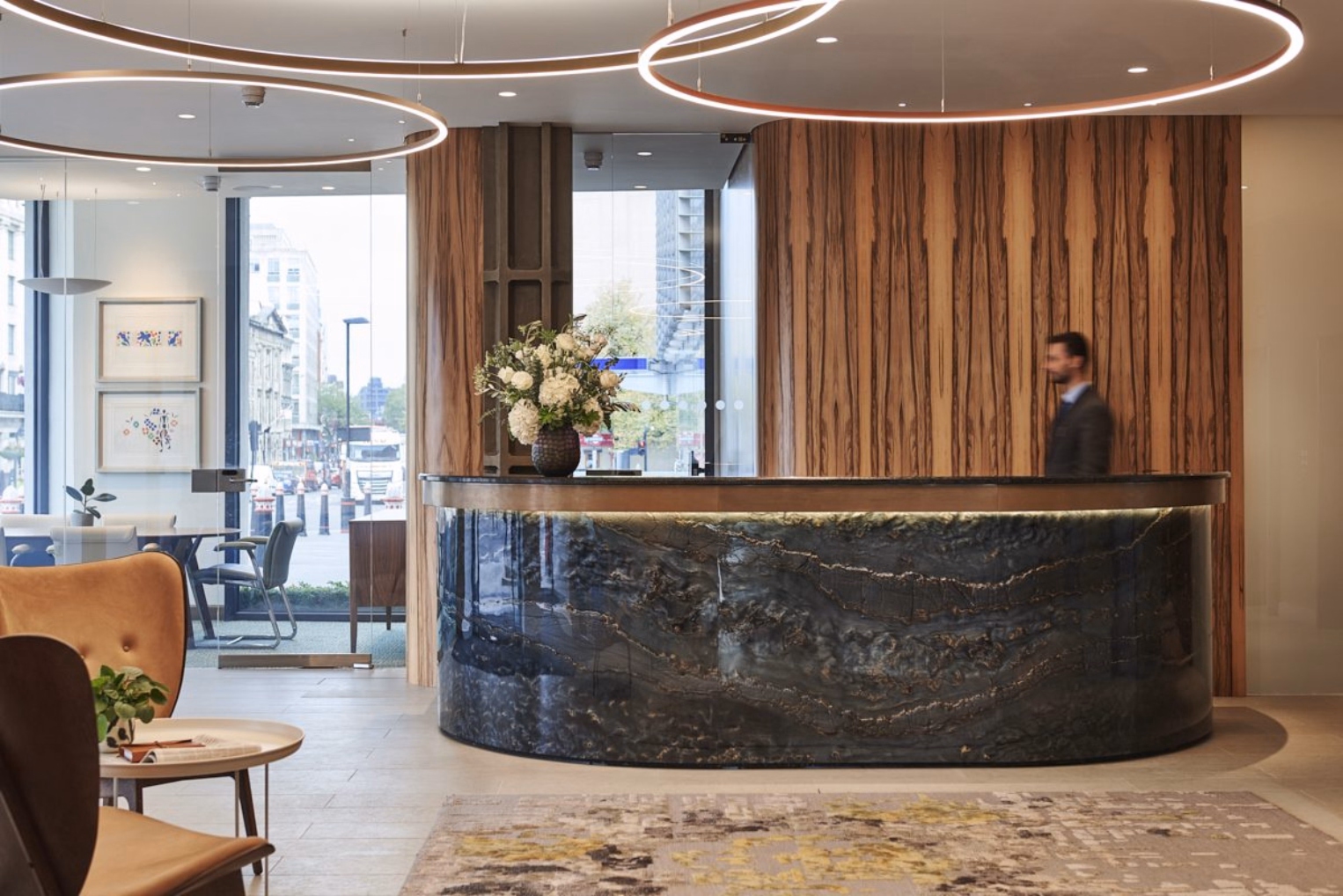 A curved, dark marble reception desk with a backlit trim in a minimalist office lobby, against a wood-panelled wall and illuminated by circular ring chandeliers.