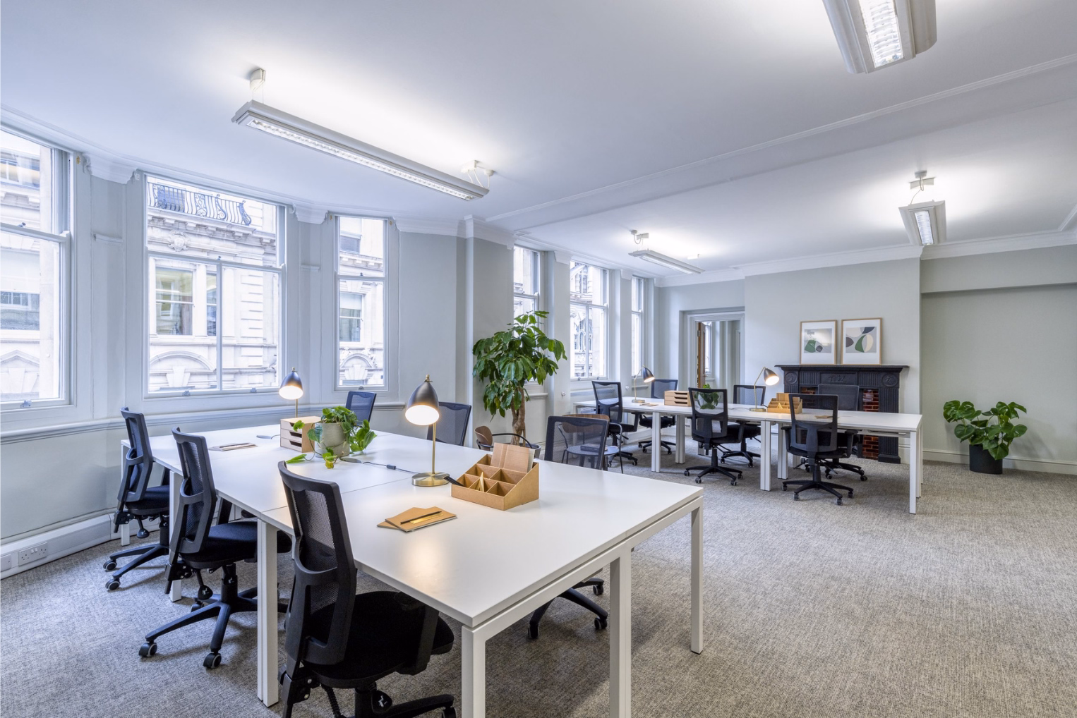 Black mesh chairs around long white desks with brass lamps with large sash windows, a fireplace mantle in the background, and leafy green potted plants.