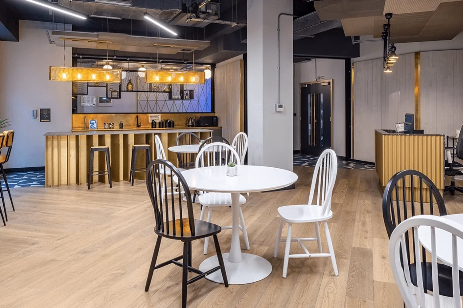 White and black chairs around round white tables and a gold-panelled kitchen bar with stools under long mesh pendant lights, and an industrial-style ceiling.
