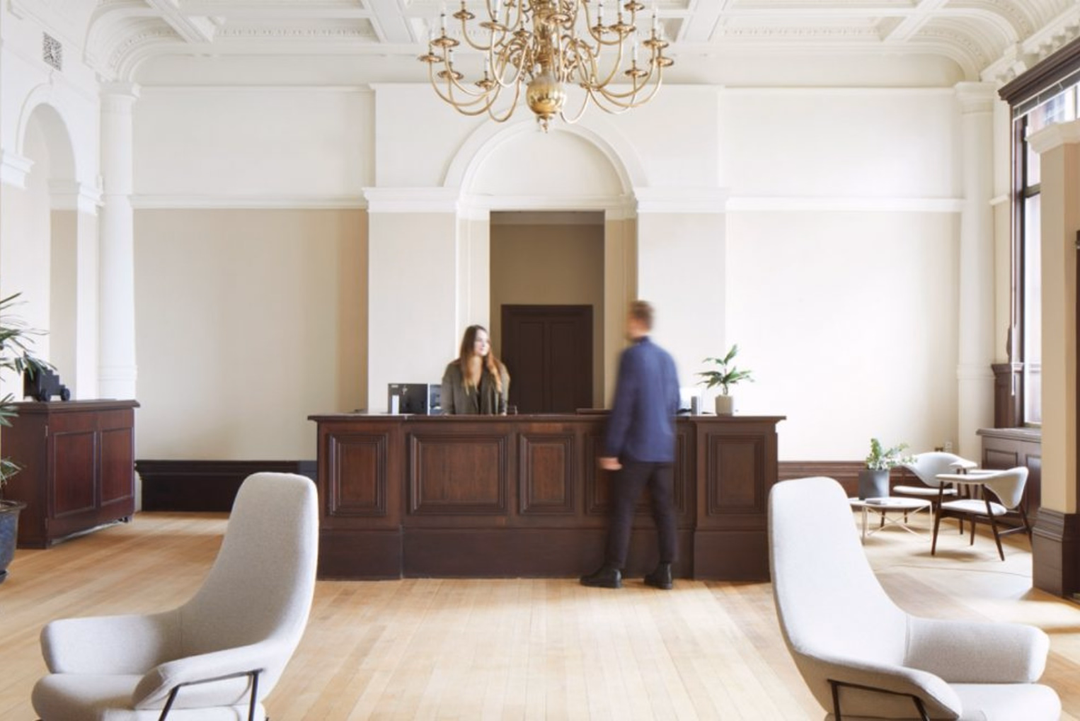 A dark wood reception desk in a high-ceiling lobby under a brass chandelier, light grey armchairs and white architectural moulding details.