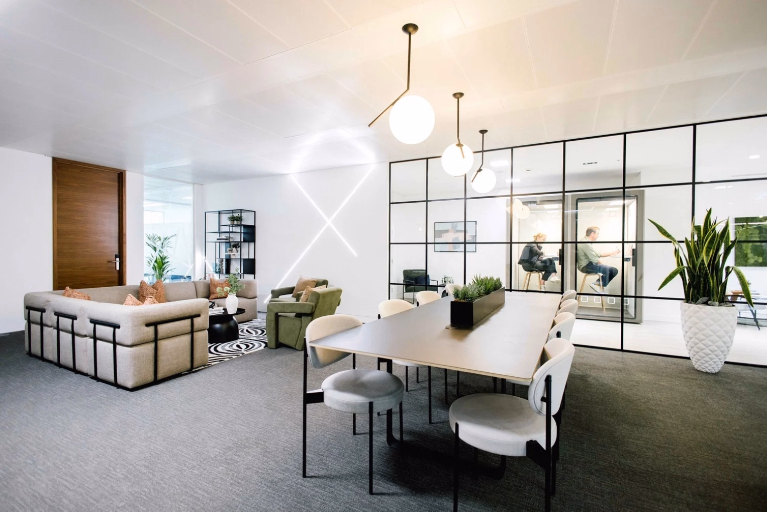 Light grey dining chairs surround a long table beneath globe pendant lights and a large grey sectional sofa with olive-green armchairs.