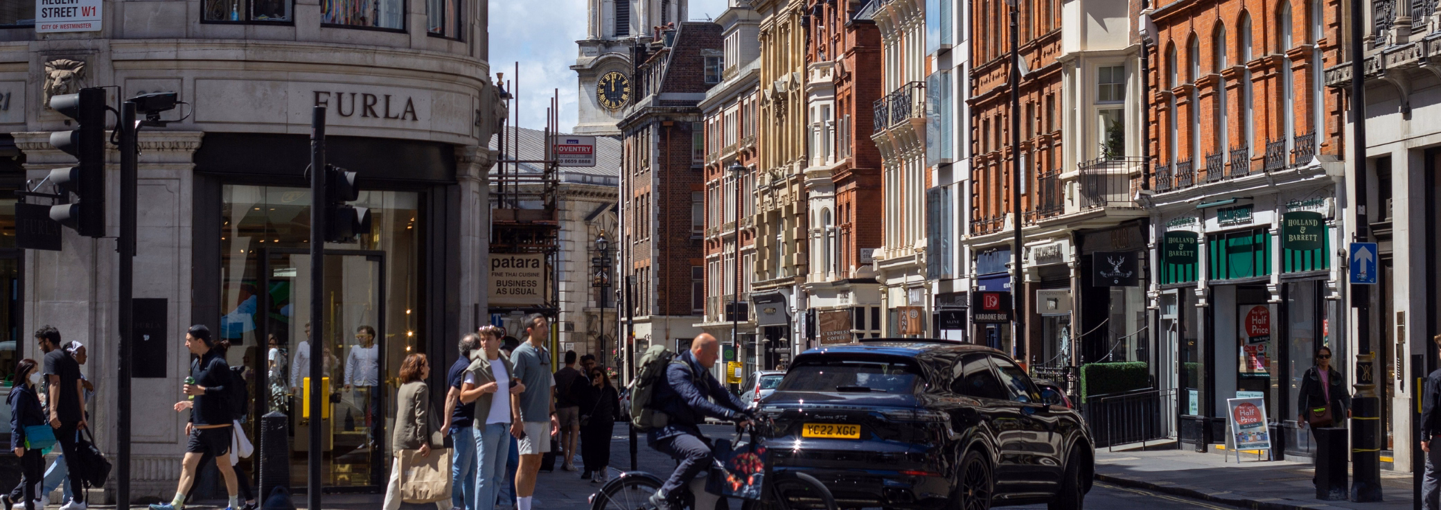 Pedestrians and a cyclist on a London street corner next to a "FURLA" storefront, flanked by a row of classic brick buildings.