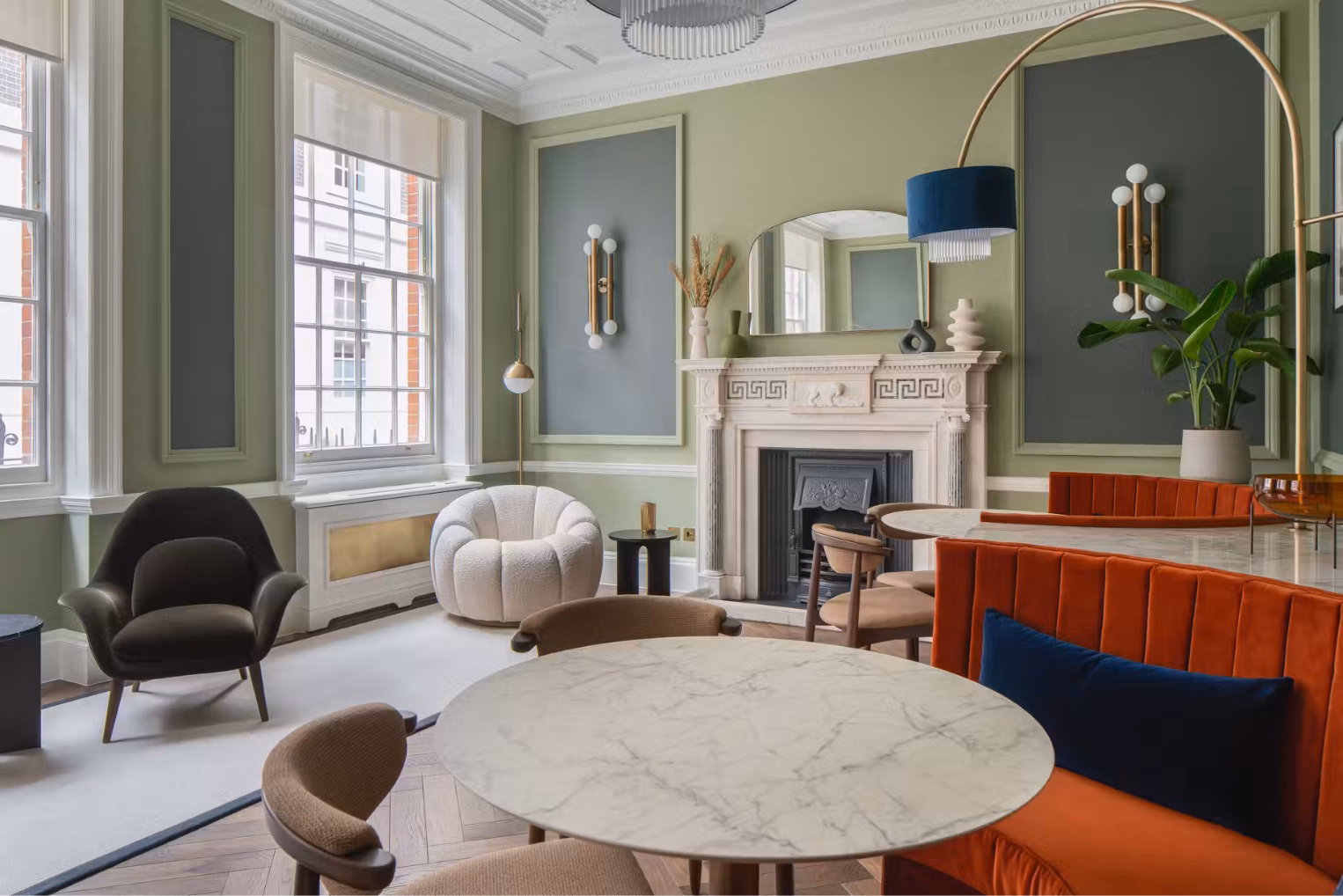 A lounge features a marble table paired with an orange banquette and cream chairs with a fireplace, a round white armchair, and sash windows in the back.