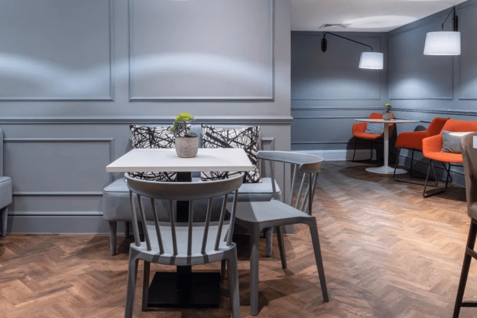 Two grey chairs at a square white table with a grey banquette bench and red armchairs in the background under wall-mounted lamps.