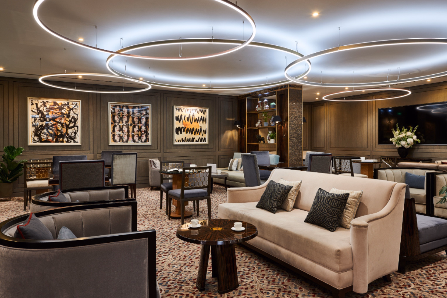 A beige sofa with patterned pillows next to grey velvet tub chairs on a patterned carpet in a room, featuring dark wood-panelled walls.